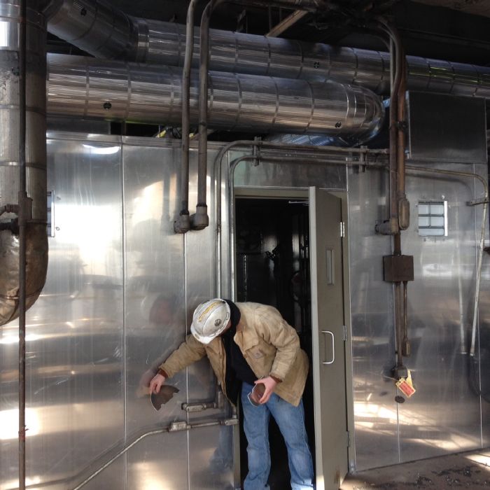 A person wearing a hard hat and work jacket examines pipes attached to a metallic industrial wall with an open door, surrounded by overhead ductwork and machinery.