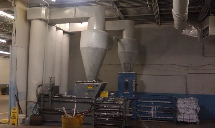 Industrial shredding and baling machine in a factory setting, with piles of shredded paper and a yellow caution sign on the floor. Large grey ventilation ducts are attached above the machinery.