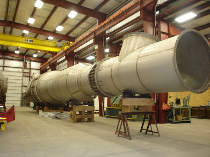 Large cylindrical metal industrial duct or pipe section supported by wooden and metal stands inside a spacious factory or warehouse with overhead crane and red steel beams visible.