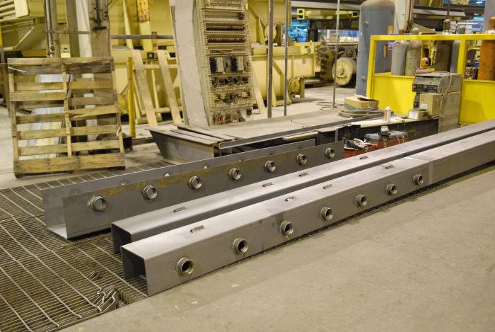 Metal industrial components with multiple circular fittings are placed on a factory floor surrounded by machinery, pallets, and equipment in a manufacturing setting.
