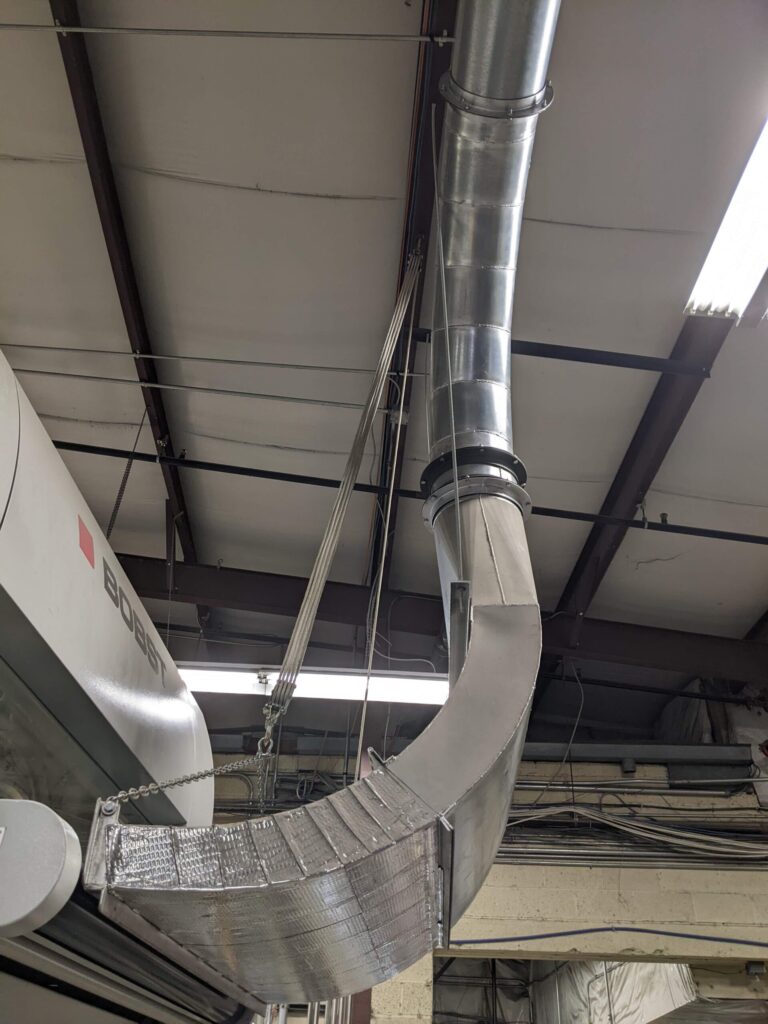 A large, silver ventilation duct—resulting from custom fabrication—curves downward from the ceiling of an industrial building, supported by metal cables. Fluorescent lights and exposed beams are visible overhead.