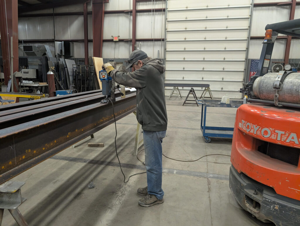 A person wearing a cap and hoodie uses a power tool on a large steel beam inside a warehouse. Nearby, an orange forklift is partially visible. The workspace has metal walls and a large overhead door.