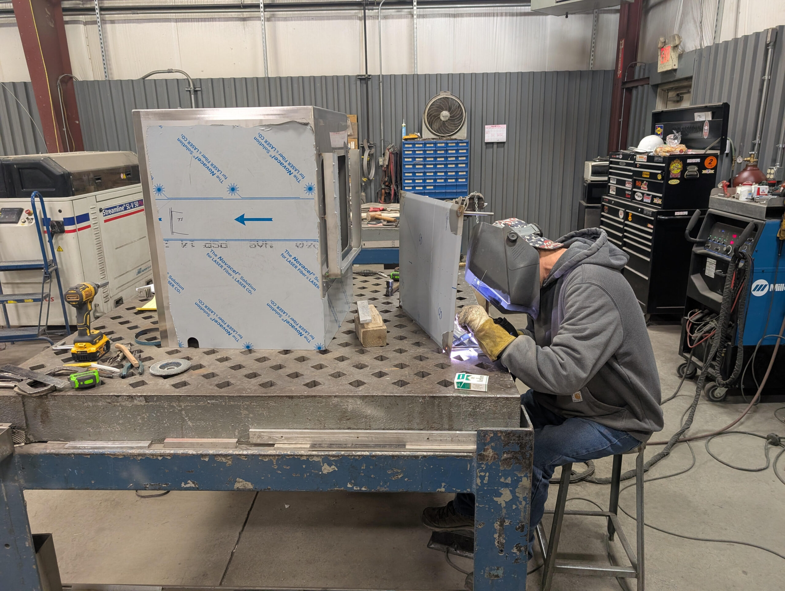 A person wearing a welding helmet and hoodie is welding metal at a workbench in an industrial workshop, surrounded by tools, metal parts, and machinery.