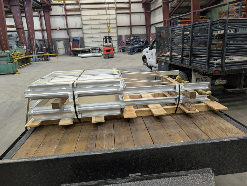Stacked metal frames are secured on a wooden pallet with straps, sitting on a flatbed trailer inside a large industrial warehouse. In the background, a white truck and various equipment are visible.