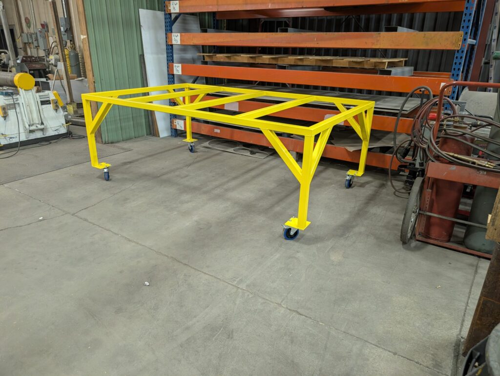 A bright yellow metal frame table on wheels is positioned in an industrial workshop, surrounded by shelves, tools, and equipment on a concrete floor.