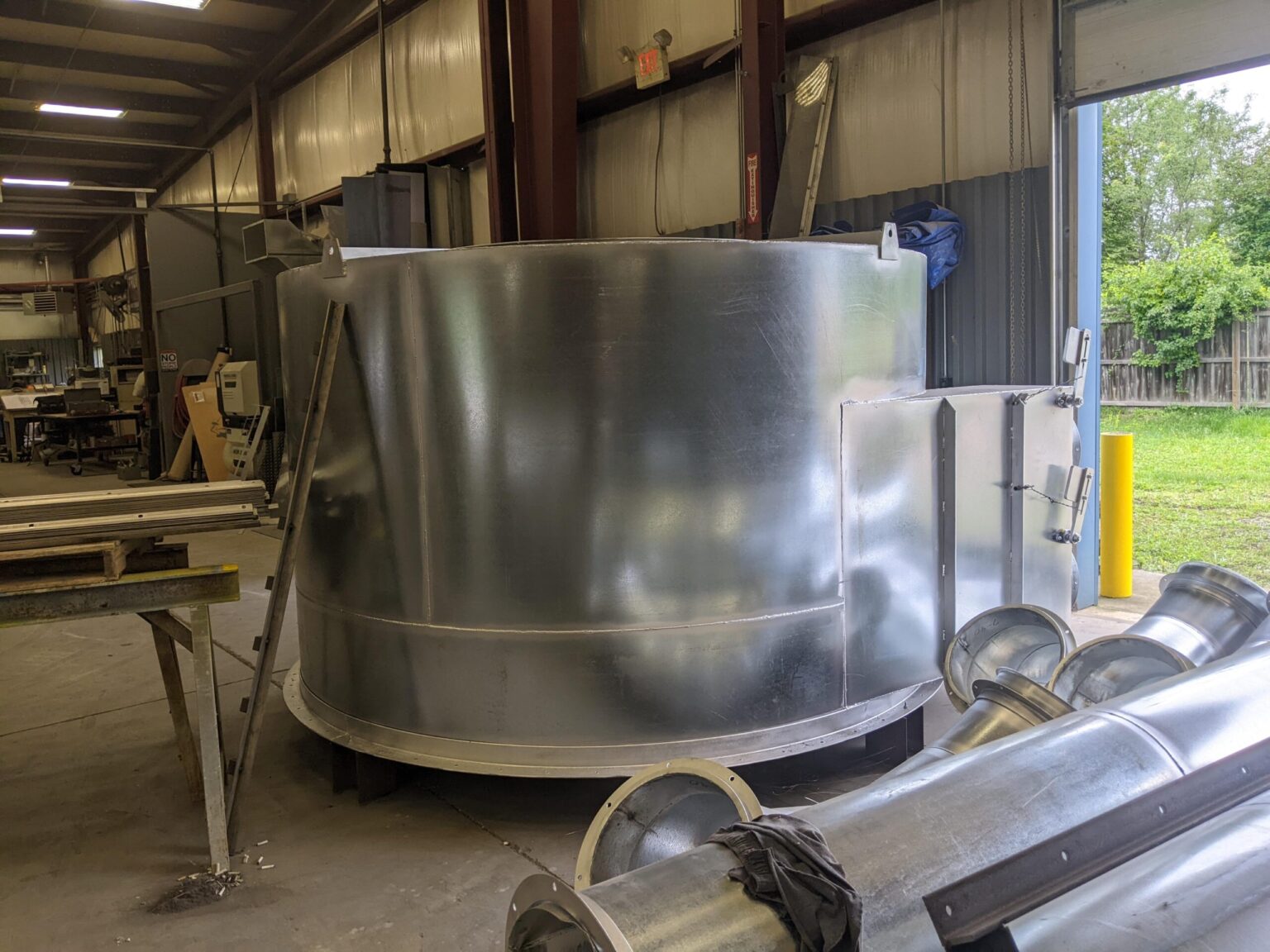 A large, cylindrical metal tank stands in an industrial workshop near an open garage door. Various metal parts and equipment are scattered around, and trees are visible outside.