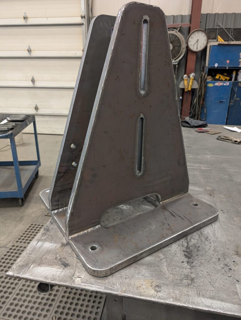 A large, triangular metal bracket with two vertical slots stands upright on a workshop table. The workshop background includes tools, a blue cabinet, a wall clock, and a partially open garage door.