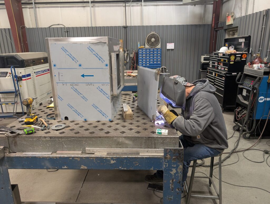 A person wearing a welding helmet operates welding equipment at a metal workstation in an industrial workshop. Metal sheets, tools, and machinery are visible on and around the table.