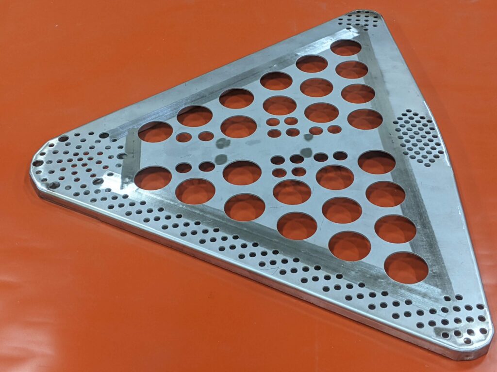 A triangular metallic plate with multiple circular holes of varying sizes arranged in patterns, placed on an orange surface. The plate has rounded corners and rows of smaller holes along the edges.