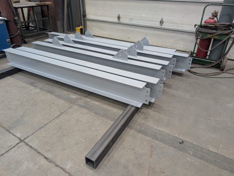 Five large, painted steel beams with bracketed ends are stacked on a workshop floor, surrounded by equipment and tools in an industrial setting.