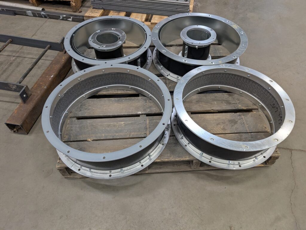 Four large metal circular duct connectors with flanges and mesh liners are arranged on a wooden pallet in an industrial setting, with concrete flooring visible in the background.