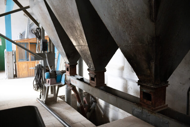 Three large, industrial metal hoppers—hallmarks of Industrial Metal Products—stand above a grated floor, connected to machinery and pipes inside a factory. A small wooden office is visible in the background.
