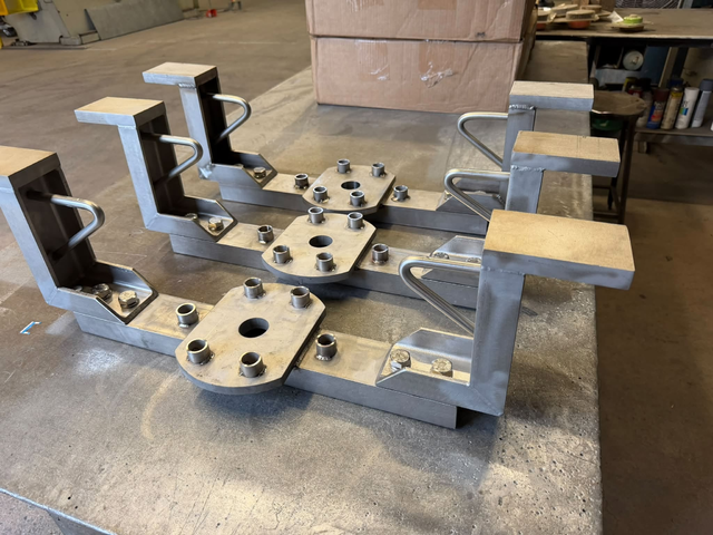 Custom-fabricated steel fixture assembly with mounting plates, bolt holes, welded brackets, and metal supports on a workshop table.